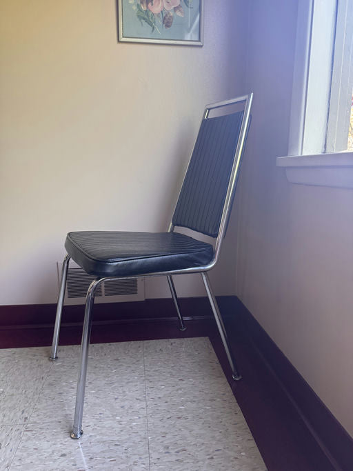 A metal and black vinyl chair is positioned in a corner of a room, beside a window and a floral artwork on the wall. The floor is tiled.