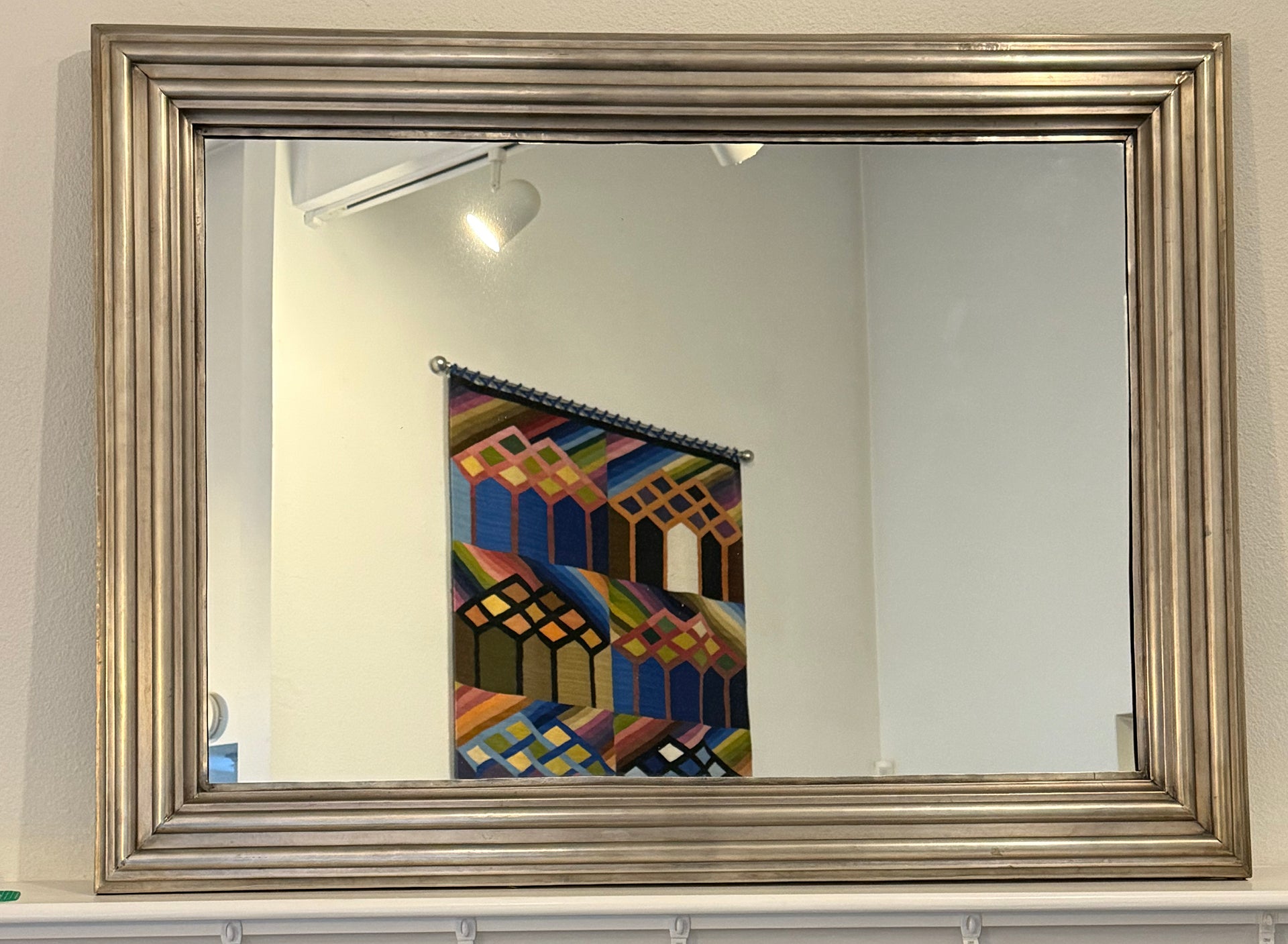 A decorative silver-framed mirror reflects a colorful wall hanging with geometric patterns, set against a light-colored wall in a bright room.