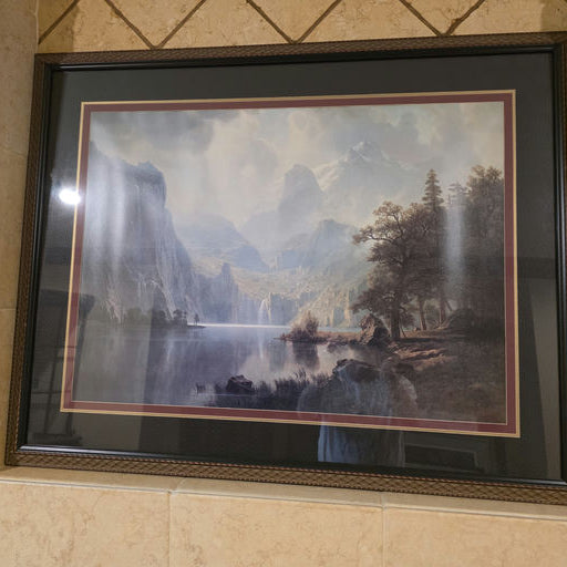 Framed landscape painting featuring mountains, a lake, and lush trees, displayed on a tiled wall. Soft colors evoke a serene atmosphere.