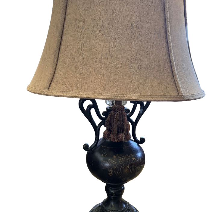 A decorative lamp with a black base and elegant cord design, topped with a beige fabric shade, sits on a patterned rug next to a bed.
