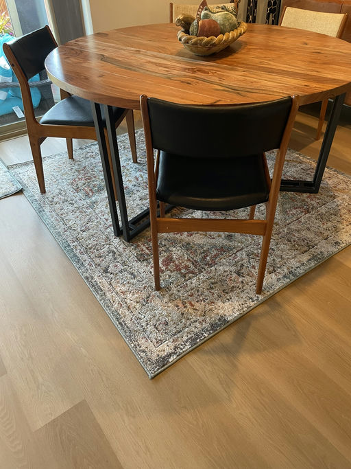 A round wooden dining table with four chairs sits on a patterned rug. A decorative bowl is placed in the center of the table.