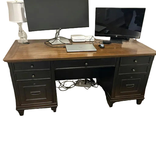 A wooden desk with two monitors, a laptop, and a lamp. The setup includes visible cables and a nightstand with drawers. The floor features laminate planks.