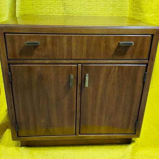 A wooden cabinet with two doors and a drawer, featuring metallic handles. The background is bright yellow, highlighting the furniture piece.