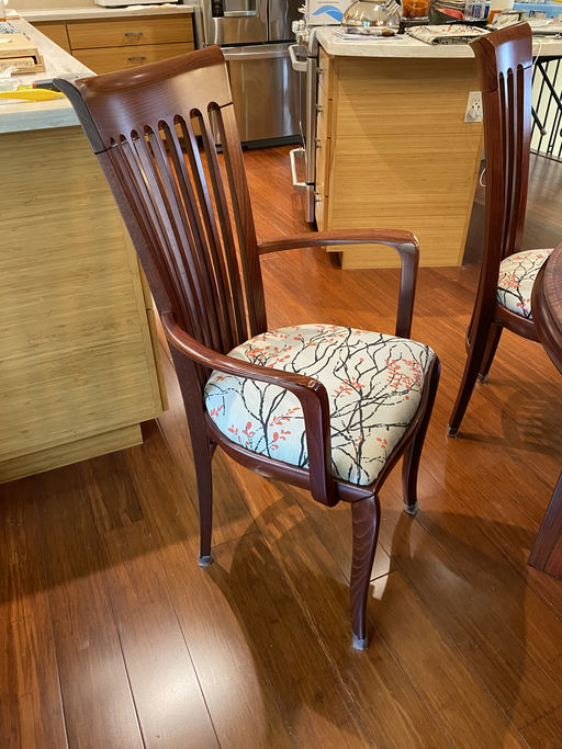 A wooden dining chair with a high back and armrests features a patterned cushion with branches and flowers. The setting includes a modern kitchen.