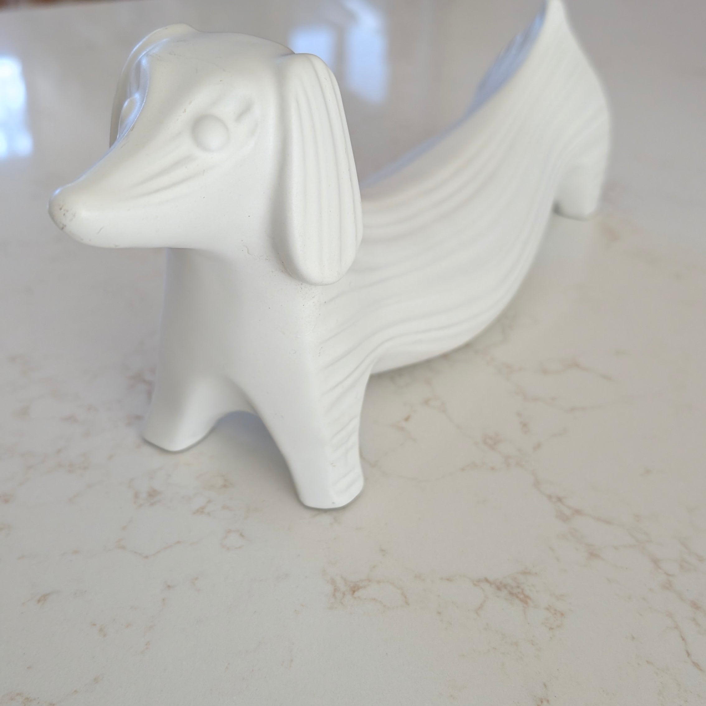 A white ceramic figurine shaped like a dachshund, with textured lines, placed on a smooth marble surface. Background includes blurred furniture.