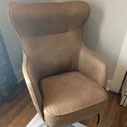 A stylish, modern armchair featuring a light brown faux leather finish and sleek black legs, positioned on a patterned rug against a wall.