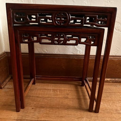 A set of two intricately carved wooden nesting tables features a rich mahogany finish and decorative openwork designs, resting on a wooden floor.