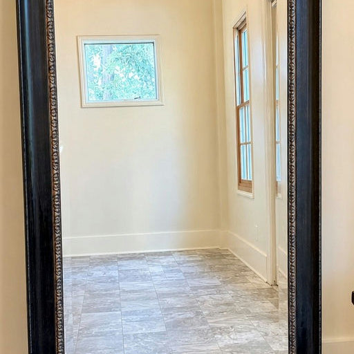 A tall, rectangular mirror with a decorative black frame reflects a bright, empty room featuring a gray tiled floor and white walls.