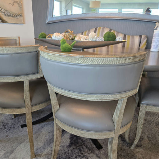 A dining area features elegant wooden chairs with gray upholstered seats, a round table, and decorative elements on the tabletop. Natural light filters in.