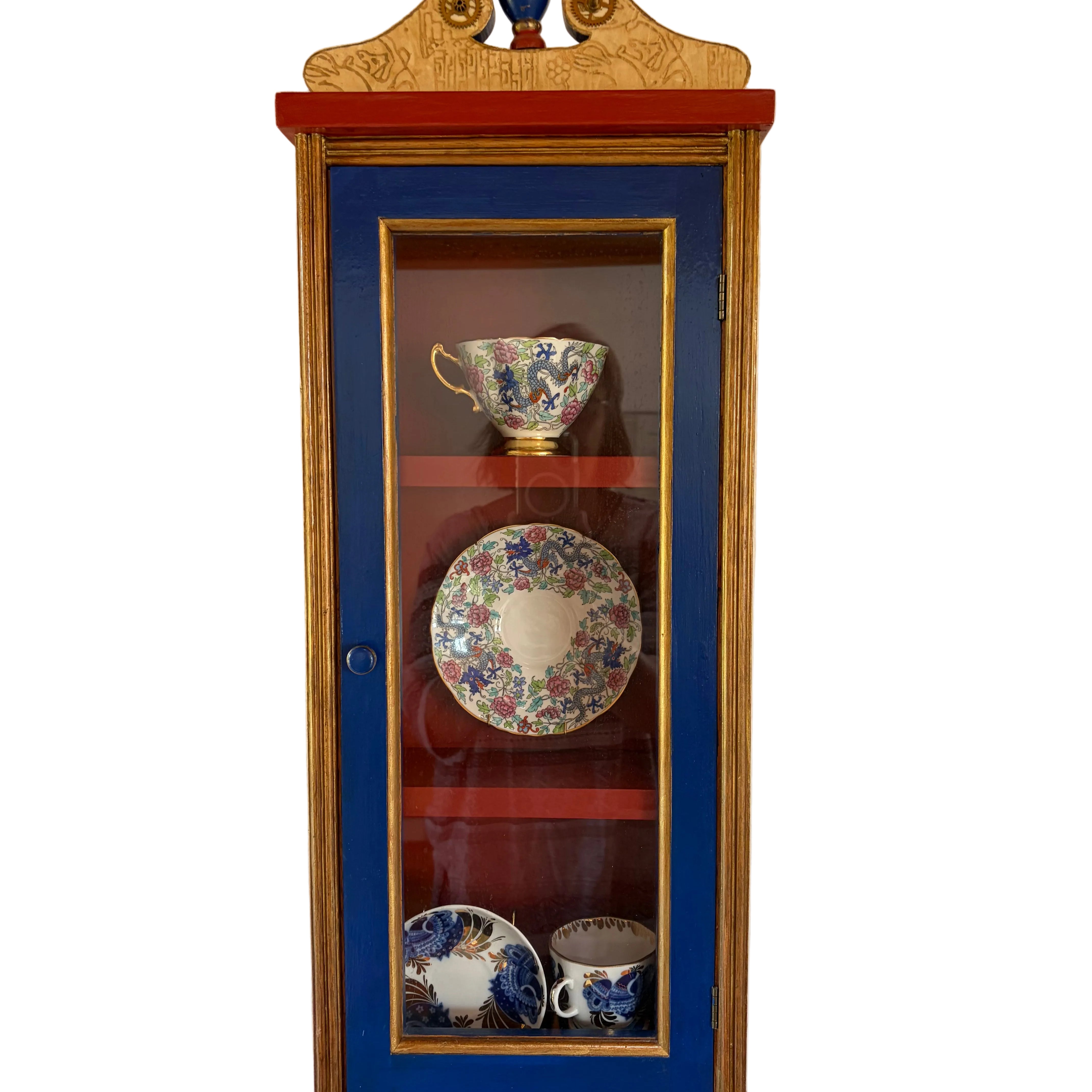 A colorful cabinet with a blue and red finish displays ornamental china, including cups and plates, against a simple white wall.