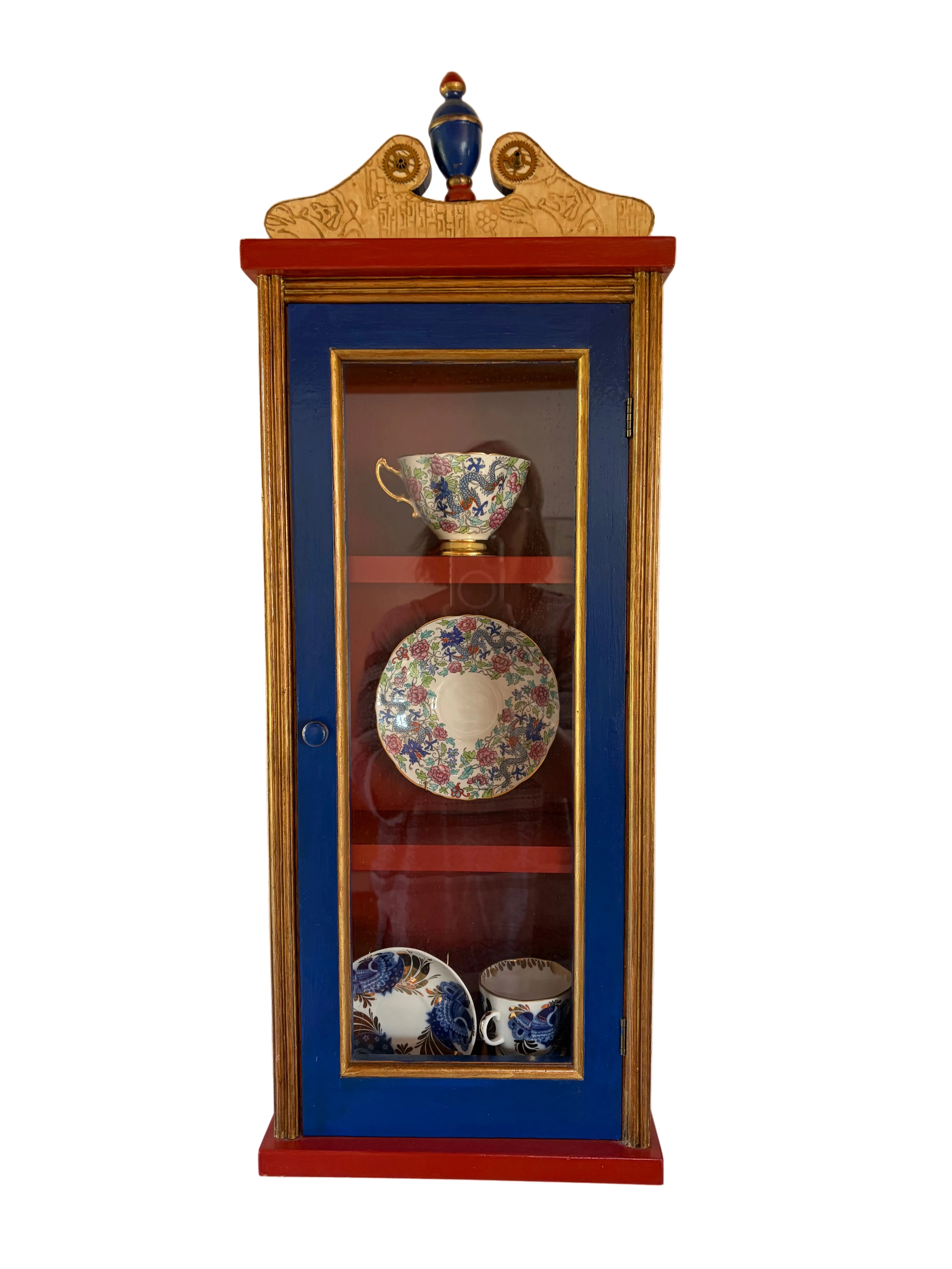 A colorful cabinet with a blue and red finish displays ornamental china, including cups and plates, against a simple white wall.