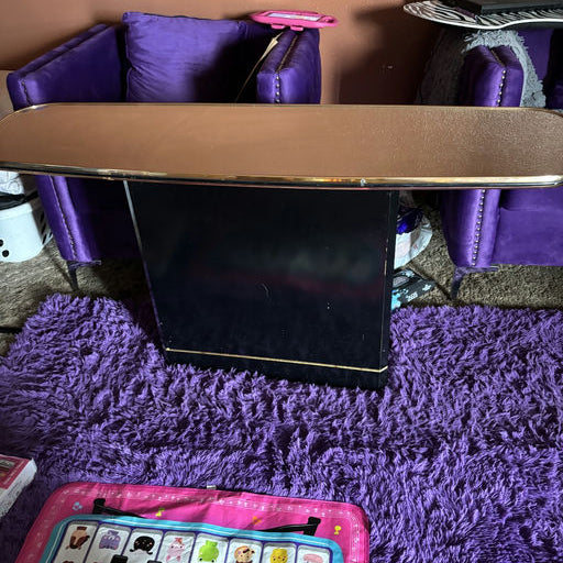 A black table with a shiny top sits in a cozy room, flanked by purple chairs and a pink play mat with colorful designs on a plush purple rug.
