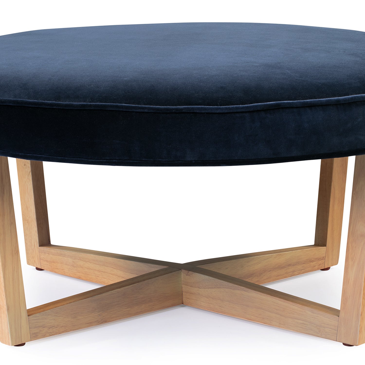 Compass ottoman has an upholstered cushioned top that is in the shape of a circle. The cushioned top has piping along the edges. There are 4 natural wood legs that angle in slightly from the top of the ottoman touch the floor. The legs connect underneath the ottoman making a shape of an X.