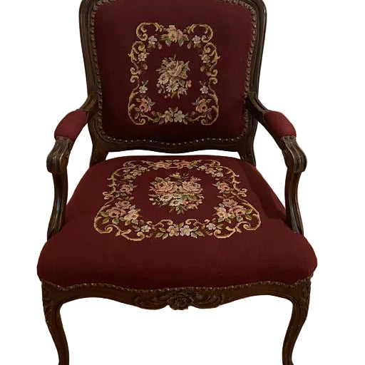 Antique armchair with ornate wooden frame and burgundy upholstery, featuring floral embroidery on the backrest and seat, placed on a wooden floor.