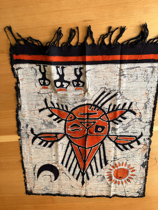 A colorful textile featuring a central red design resembling a sun or mask, surrounded by intricate patterns, with black and orange detailing.