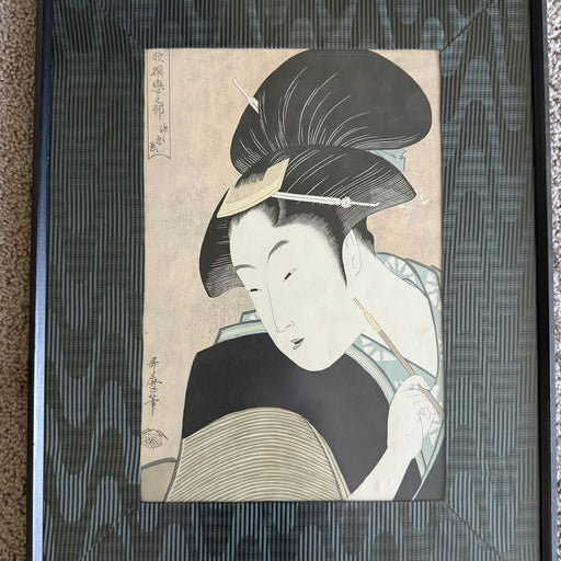 A framed ukiyo-e print features a woman with elaborate black hair and traditional clothing, holding a brush and gazing thoughtfully.