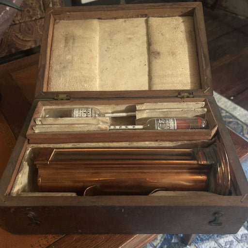 A vintage wooden box is open, displaying a copper instrument, glass vials, and other components, all resting on a felt-lined interior.