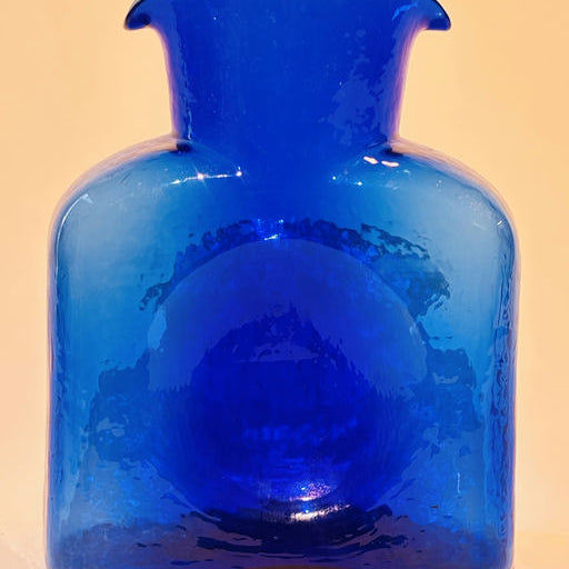 A vibrant blue glass vase with a unique shape, featuring a wide body and a narrow spout, placed against a soft, neutral background.