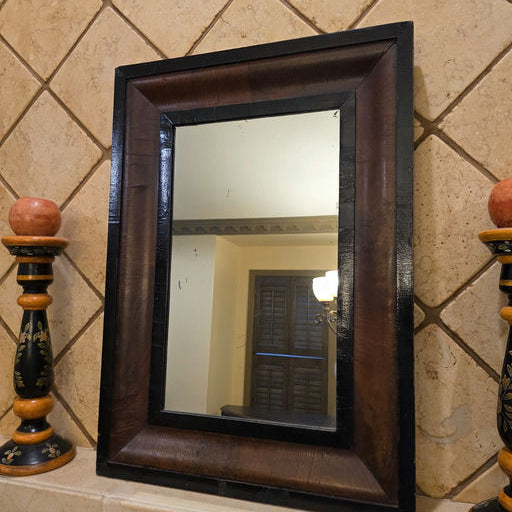 A rectangular mirror bordered with dark wood is mounted on a tiled wall. Two decorative candlesticks are positioned on either side of the mirror.