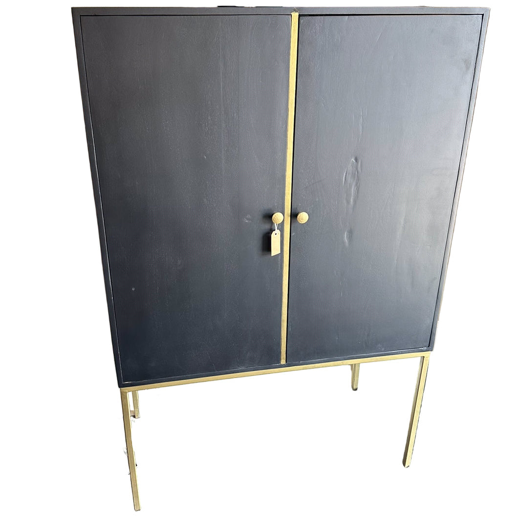 A black cabinet with gold accents, featuring two doors and round knobs, stands on a gold metal frame. A lamp and small plants are visible nearby.