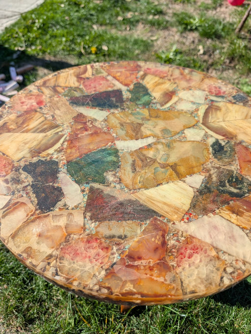 A close-up of a colorful mosaic table, featuring various polished stones in hues of orange, red, green, and tan, set against green grass.