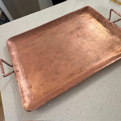 A large, rectangular copper serving tray with hammered texture, featuring two handles, resting on a speckled countertop.