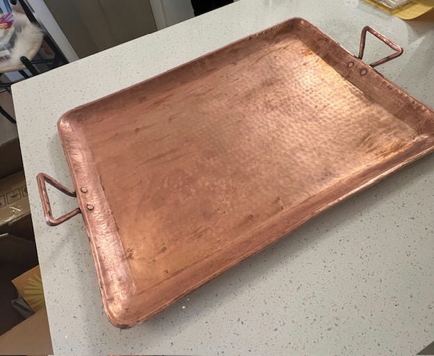 A large, rectangular copper serving tray with hammered texture, featuring two handles, resting on a speckled countertop.