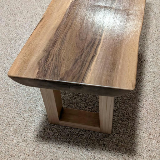 A wooden bench with a curved top showcasing rich grain patterns, supported by a simple rectangular base, set against a textured carpet background.