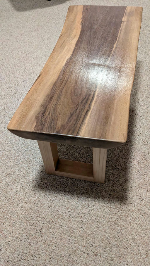 A wooden bench with a curved top showcasing rich grain patterns, supported by a simple rectangular base, set against a textured carpet background.