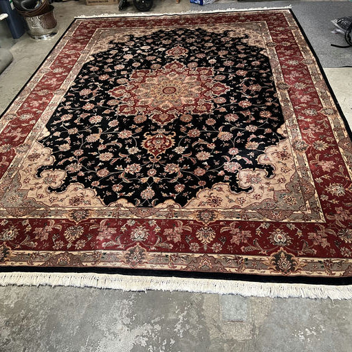 A large, intricate Persian rug featuring a floral pattern in rich black, red, and gold tones, laid on a concrete floor.