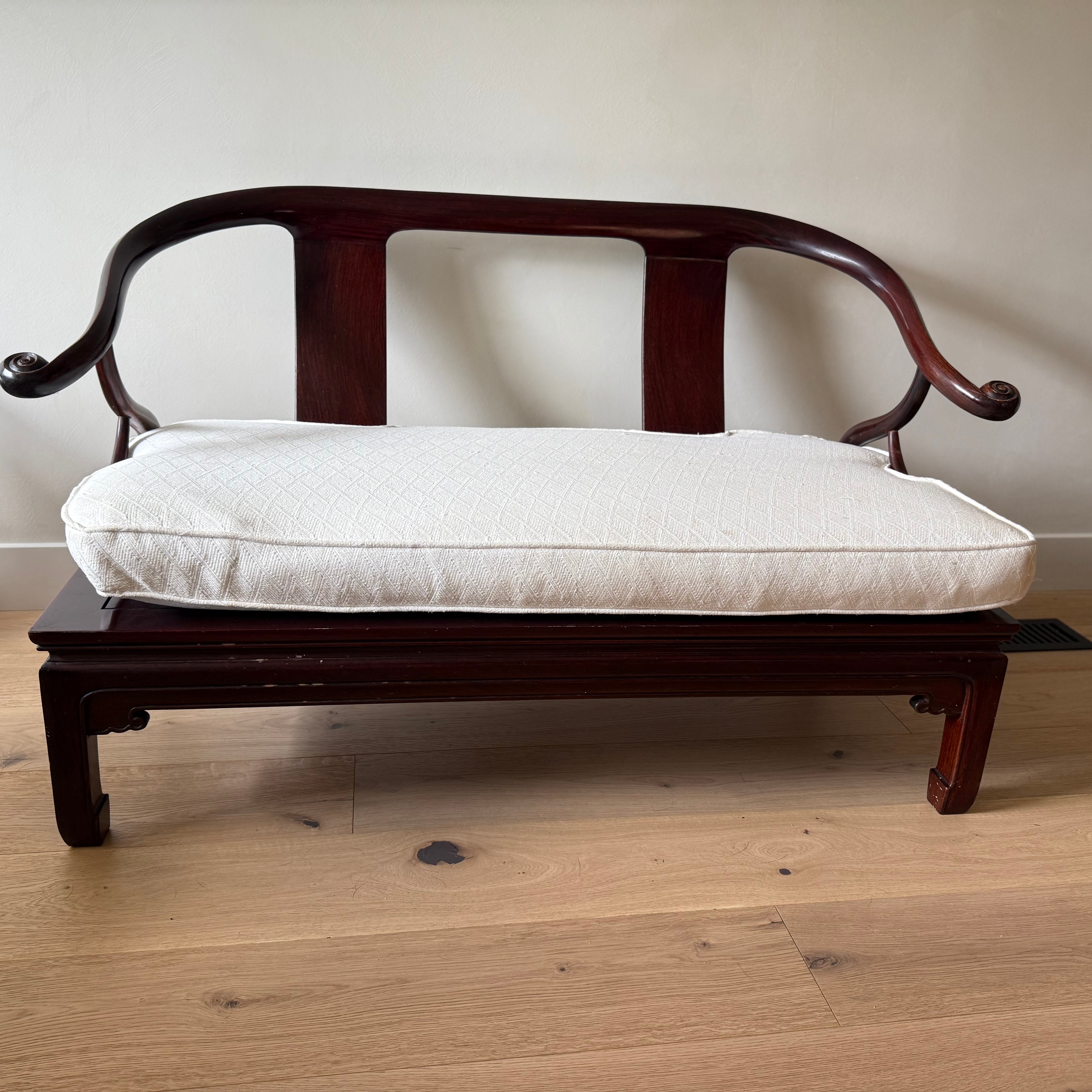 A small, elegant wooden bench with curved arms and a cream-colored cushion sits on a light wooden floor against a plain wall.