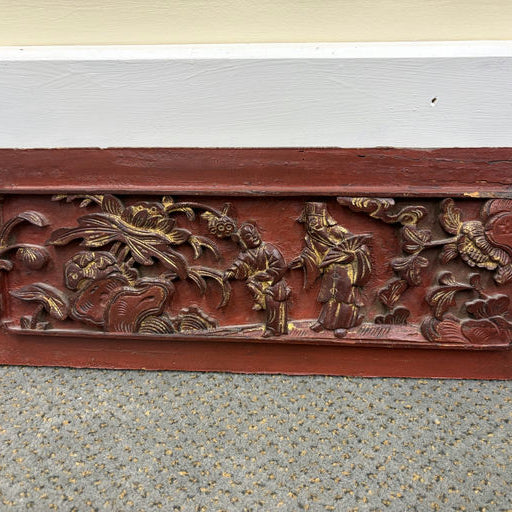 A decorative wooden panel featuring intricate relief carvings of flowers and figures, painted in red and gold, resting on a carpeted floor.