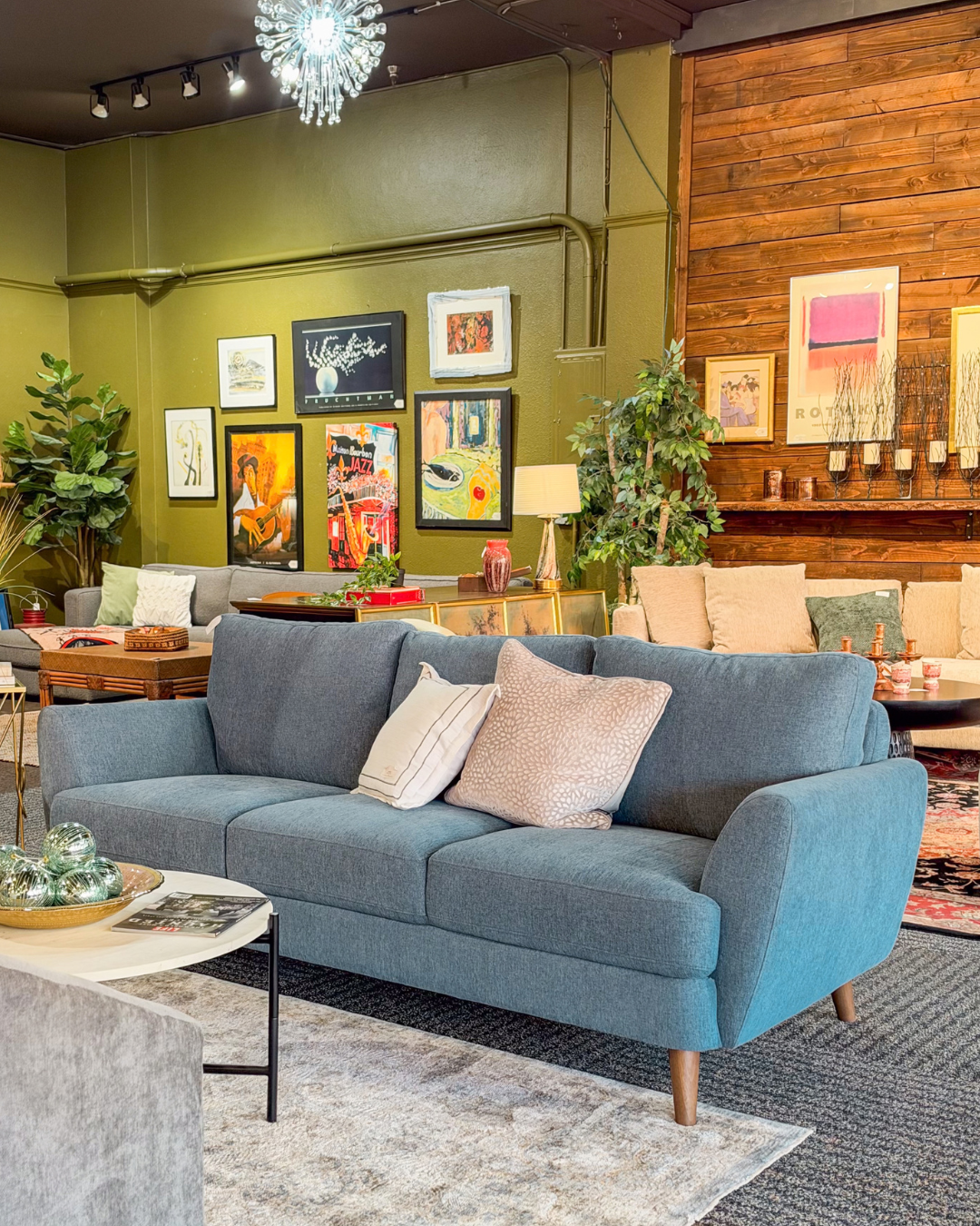 A blue upholstered sofa with light beige and white throw pillows, positioned in a styled living room showroom with a wooden wall, framed art, and other furniture in the background.