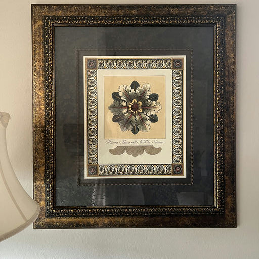 A framed botanical print featuring a floral design, accented with ornate borders and a decorative background, hung on a wall near a lamp.