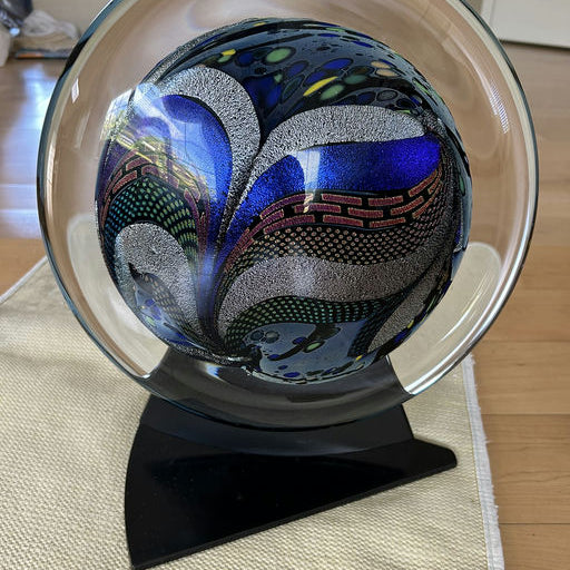 A colorful glass orb featuring intricate patterns of blue, green, and metallic accents, displayed on a sleek black stand.
