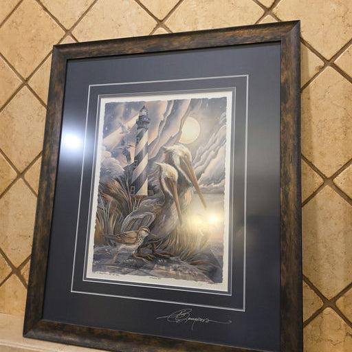 A framed artwork depicting a lighthouse, pelicans, and other birds amidst a dramatic sky, hanging on a textured tiled wall.