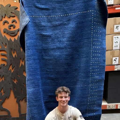 A smiling young man sits in front of a large blue fabric, next to a playful wooden figure resembling a furry creature, with shelves of boxes behind.