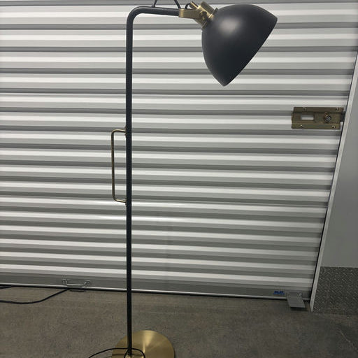 A modern floor lamp featuring a black shade and a brass accent, set against a backdrop of corrugated metal storage unit doors.