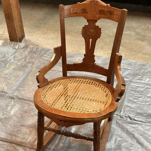 A vintage wooden chair with a woven cane seat, featuring ornate carvings on the backrest. It's placed on a tarp in a garage setting.