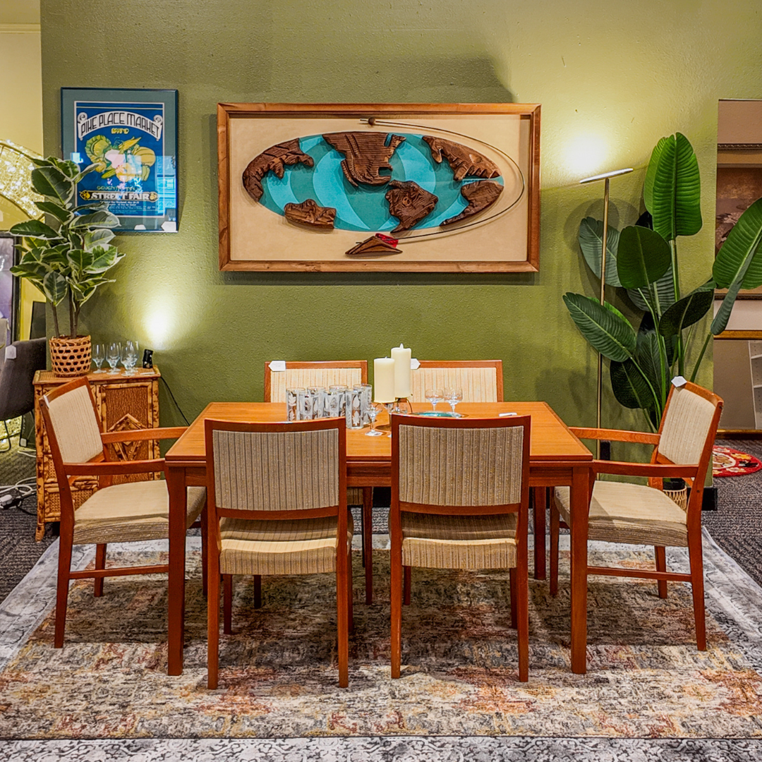 A wooden dining table with six woven-back chairs is arranged on a patterned area rug in a styled room with olive green walls, wall art, plants, and warm ambient lighting.