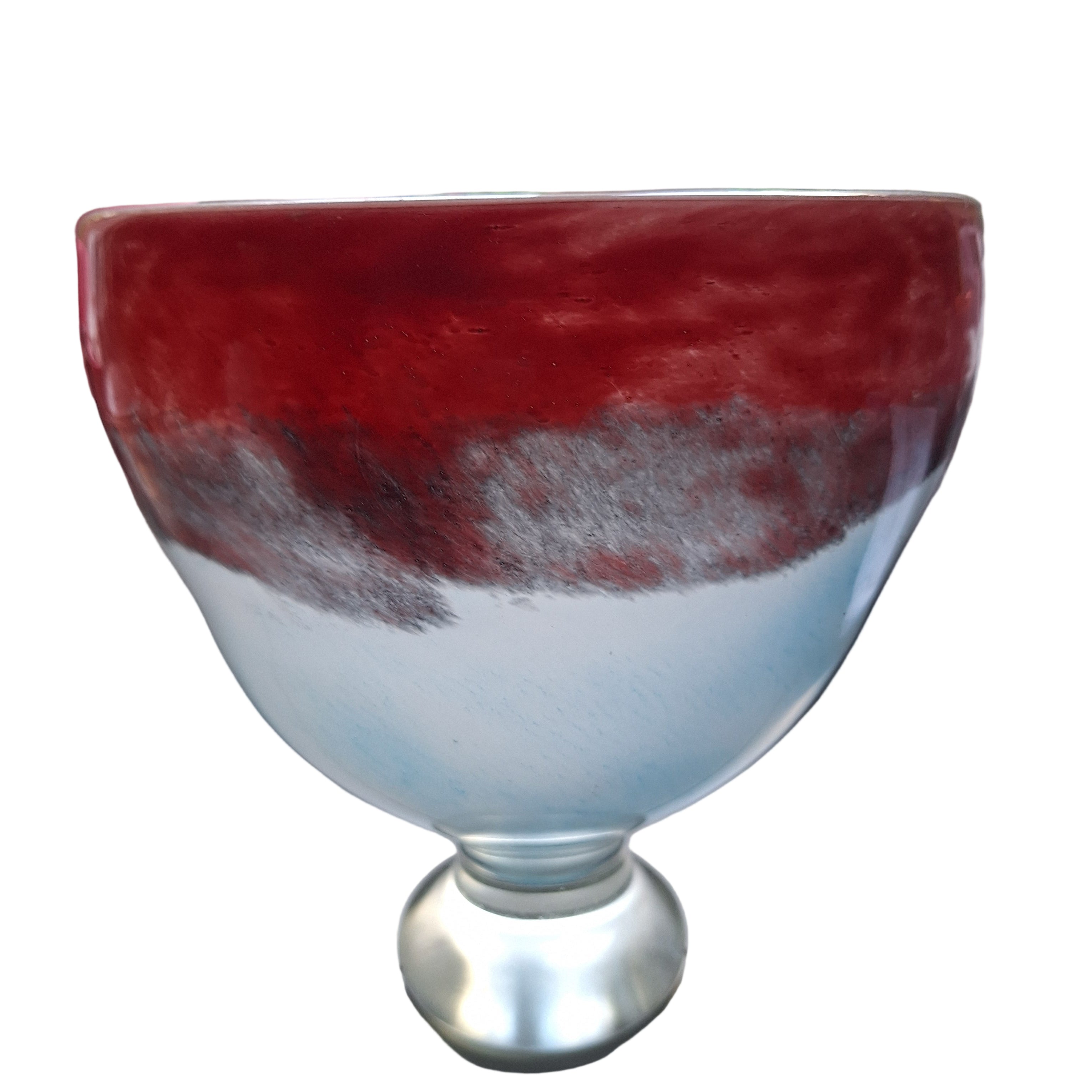 A round, decorative glass bowl featuring a red and blue gradient design, resting on a light-colored surface near a window with green foliage visible.