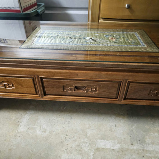 A wooden coffee table with a glass top and decorative inlay design. It features two drawers with unique handles, placed on a concrete floor.