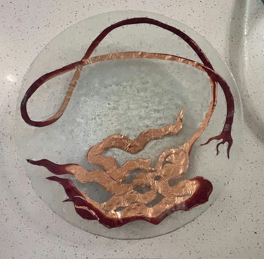 A circular glass table featuring intricate copper artwork resembling flowing waves and abstract shapes, set against a light speckled surface.