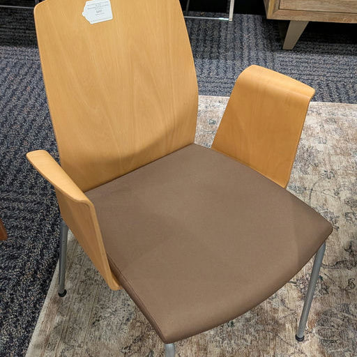 A modern chair features a light wood back, brown seat cushion, and metal legs. It has armrests and is set against a patterned rug and green stools.