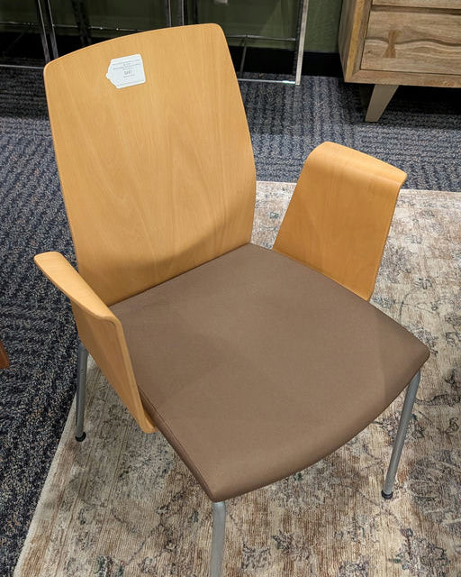 A modern chair features a light wood back, brown seat cushion, and metal legs. It has armrests and is set against a patterned rug and green stools.