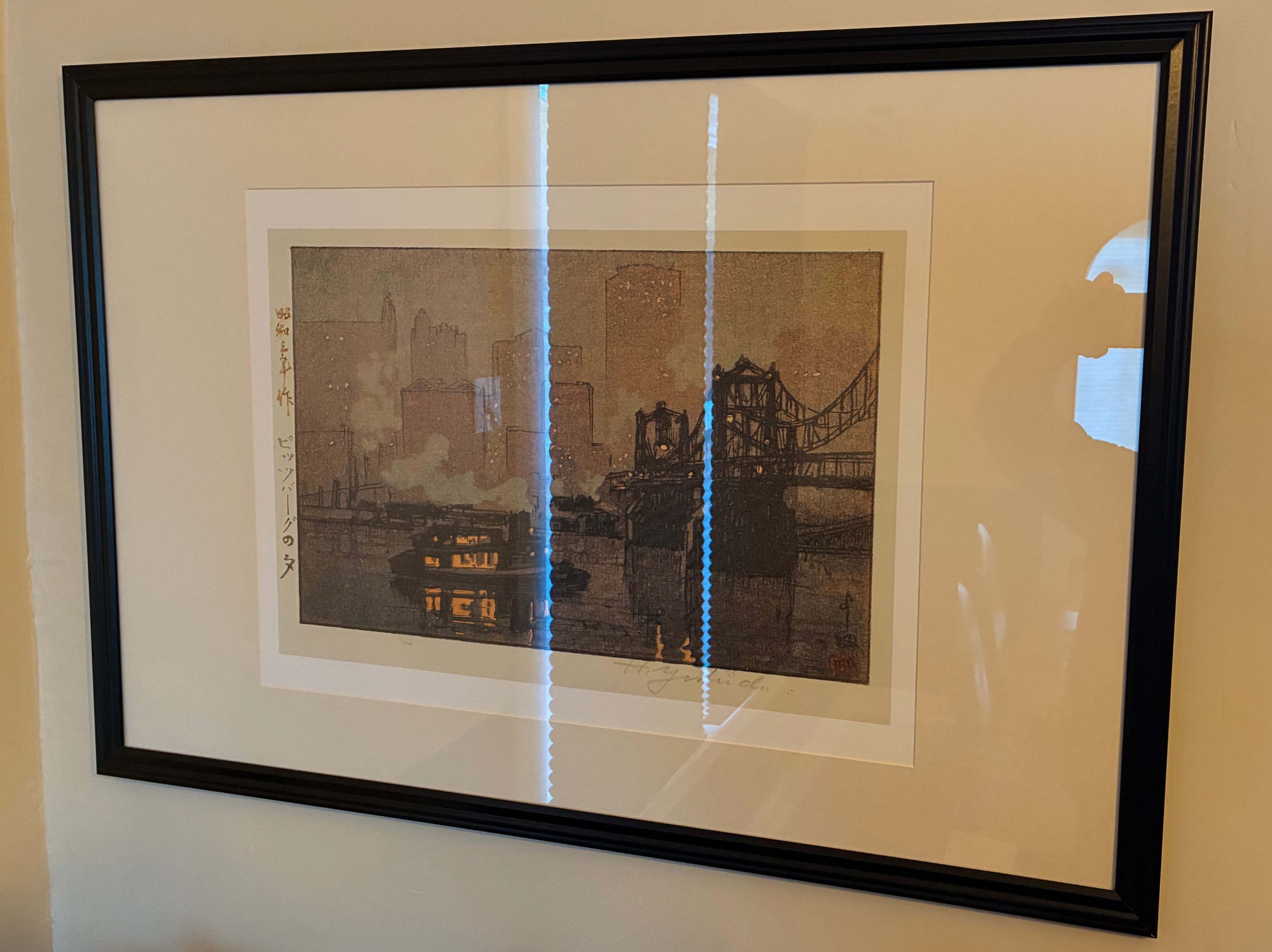 Framed artwork featuring a cityscape with a bridge, smoky atmosphere, and a boat on water, hung on a wall in a brightly lit room.