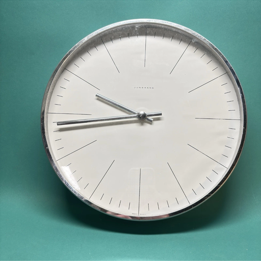 A round wall clock with a minimalist design, featuring a white face, subtle markings, and silver hands, set against a teal background.