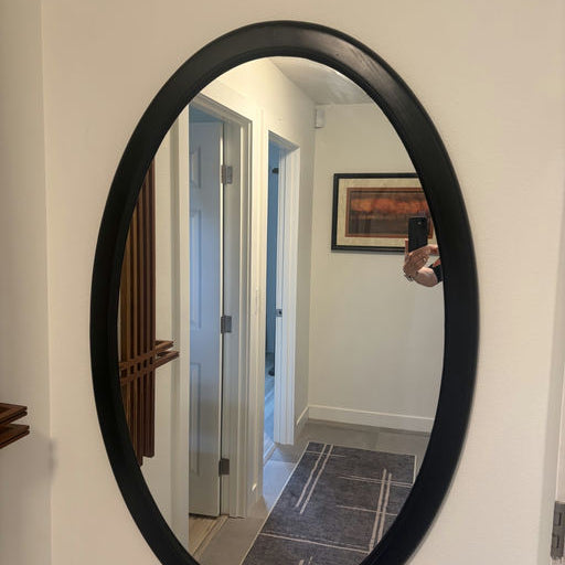 An oval mirror with a black frame reflects a well-lit hallway and a patterned rug, showcasing a bright, modern interior design.