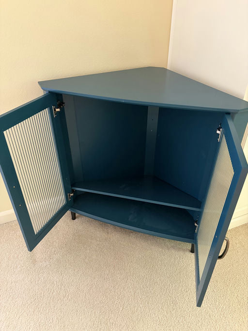 A teal corner cabinet with a triangular shape and doors revealing interior shelving. The cabinet is placed on a soft carpet against a wall.