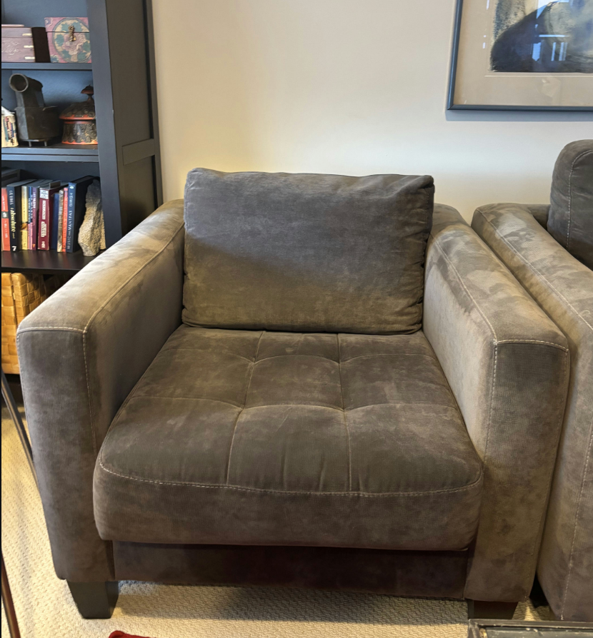 A gray upholstered armchair with a tufted seat cushion, situated in a cozy room with shelves filled with books and decorative items in the background.